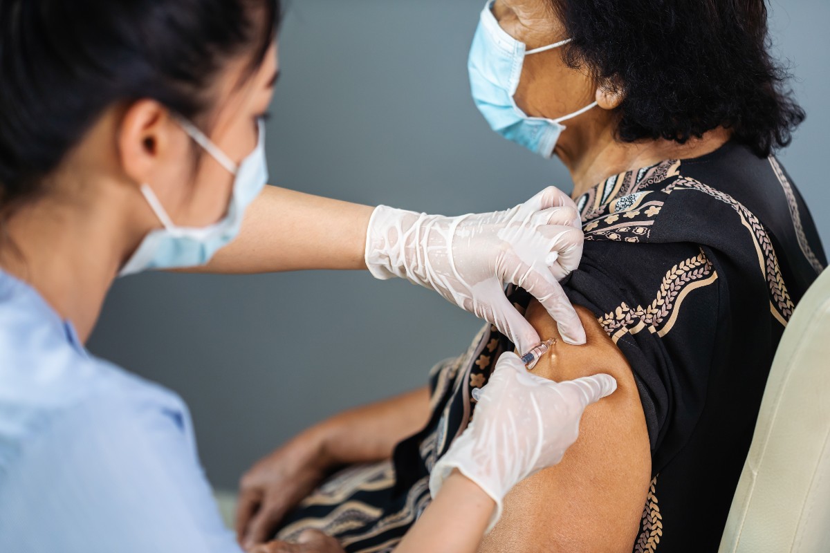 Doctor in gloves holding syringe and providing vaccine injection to senior patient in medical mask.